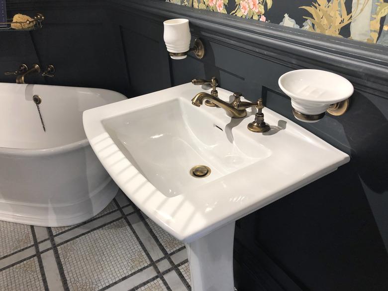 Image of luxury washroom bathroom with large freestanding bath with curved ends and bronze mixer tap, white rectangular sink wash basin with soap dish, wood panelling painted black and floral wallpaper with flowers pattern, mosaic tiled flooring tiles