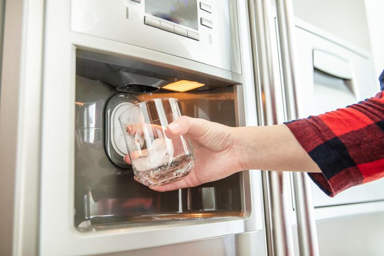 Woman's hand holds glass and uses refrigerator to make fresh clean ice cubes.