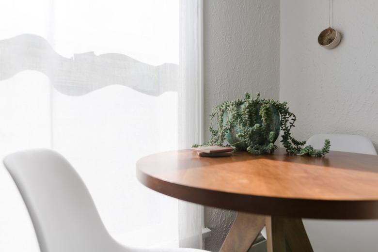 A round wood table with a succulent plant and small coasters. A curved midcentury style chair is beside and a curtained window.