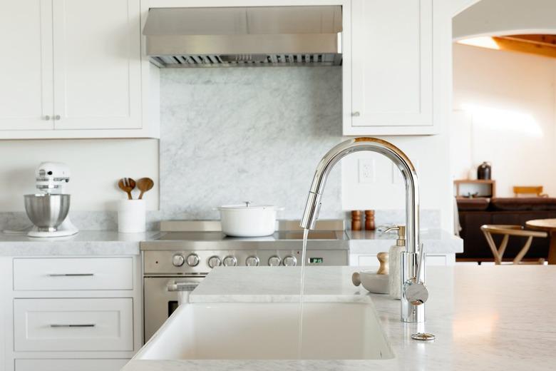 In a kitchen, a sink in a marble countertop, part of a kitchen island. The chrome faucet is running water. In the background, there's a chrome range with a white dutch oven on top.