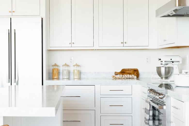 Kitchen counter. The countertop itself is marble, and the cabinets above and below are white. On the counter, three glass food storage containers, a cutting board, a string of garlic, and a white KitchenAid stand mixer with a stainless steel bowl.