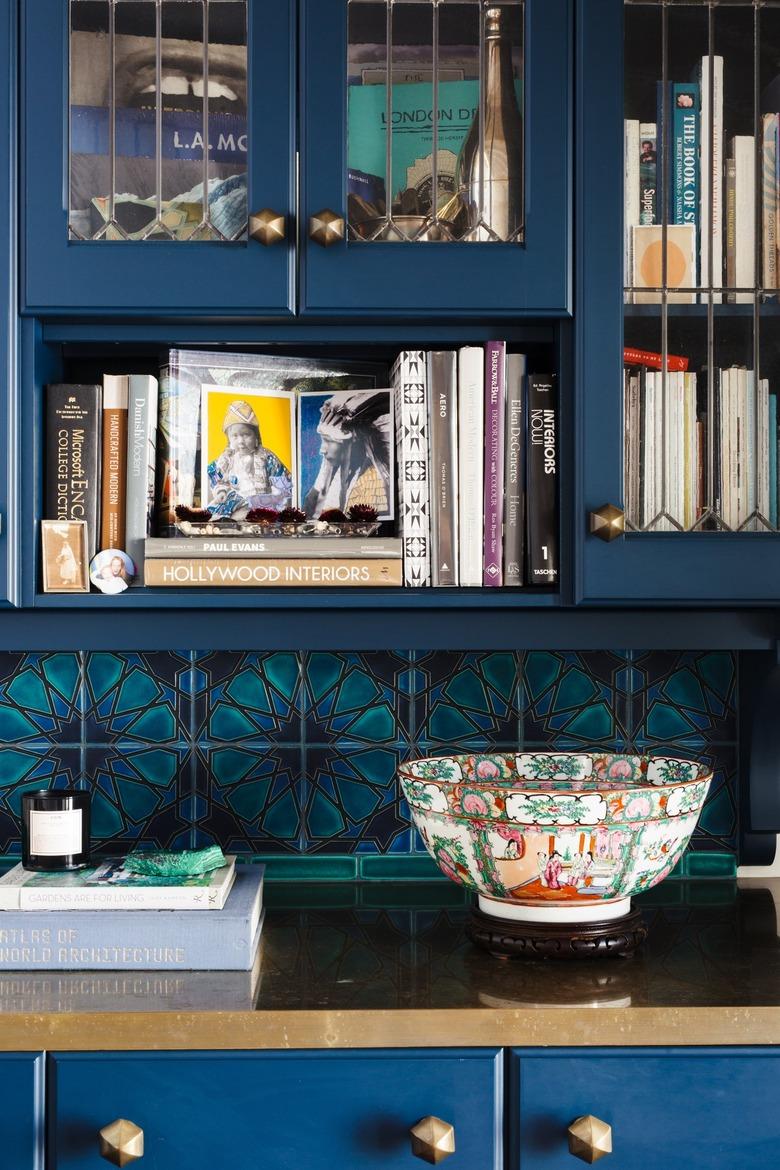 Dark blue kitchen cabinet with gold counter and knobs. Turquoise geometric floral backsplash, vintage bowl and books.