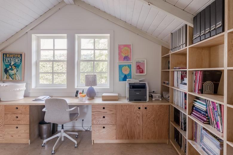 An office with a wood bookcase and Scandinavian style wood desk. A gray office chair and gray table lamp. The desk is by a window.