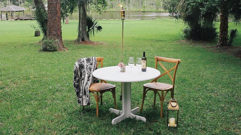 A small table and chairs with a bottle of wine and two glasses; a green lawn with trees and a river in the background