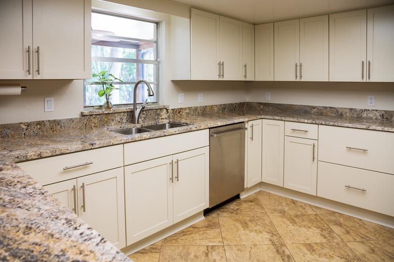 A modern U-shaped kitchen with granite countertops, white cabinets, and a touchless faucet with double-bowl stainless steel sink in front of a window.