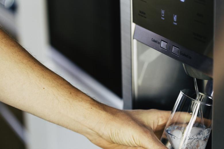 Male hand is pouring cold water and ice cubes from dispenser of home fridge.