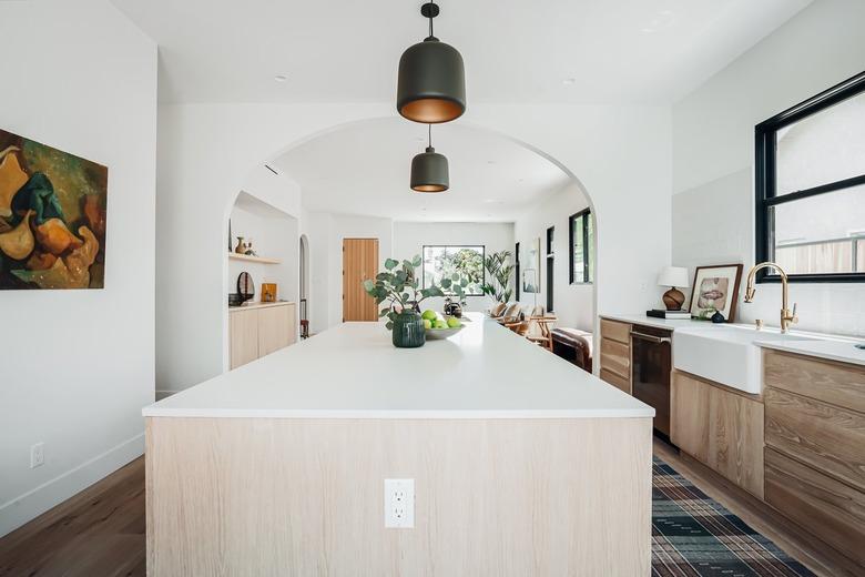 Large white and wood kitchen island with black pendant lights, farmhouse sink, and brass faucet, plaid floor rug. Black window frames.