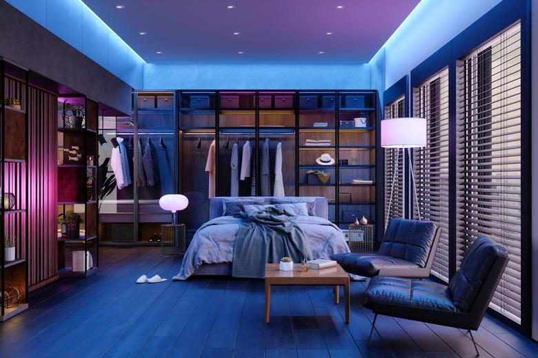 Modern Bedroom Interior At Night With Neon Light. Messy Bed, Clothes In Closet, Armchairs And Floor Lamp.