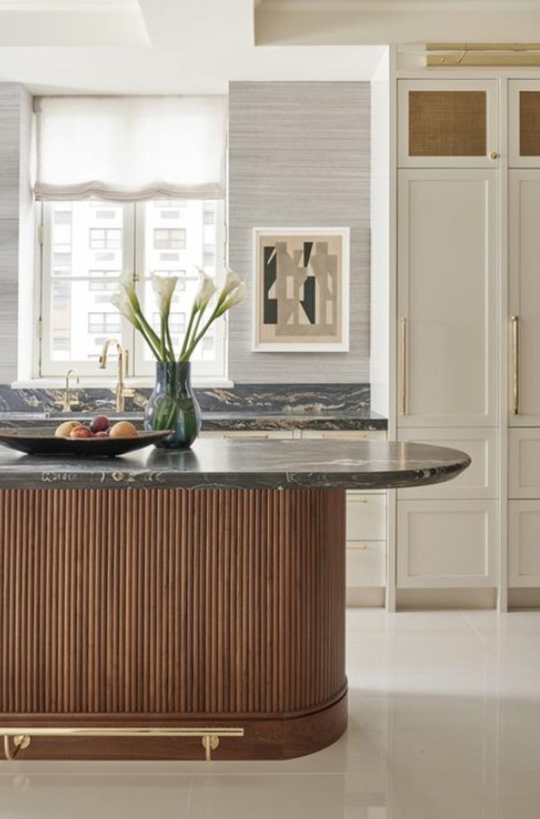 Oval kitchen island with dark gray marble countertop, wood base and brass hardware.