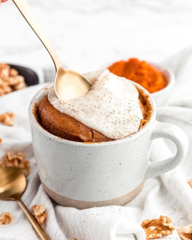 A pumpkin mug cake topped with white cream in a white mug on a white cloth.