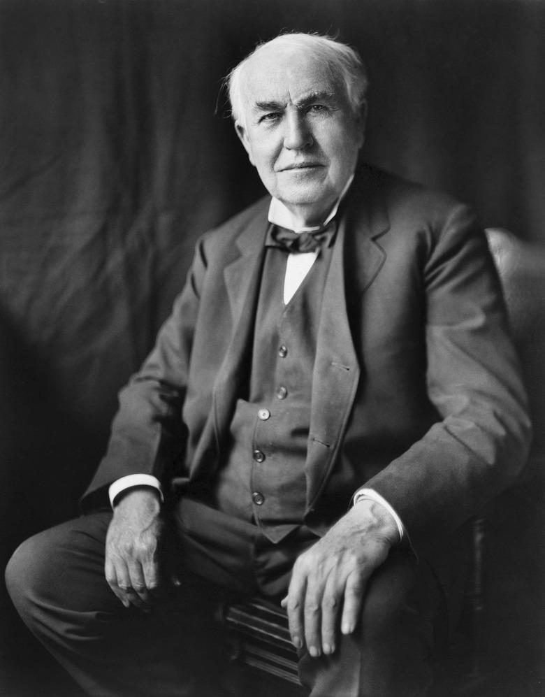 A black and white portrait of Thomas Edison sitting in a chair with his hands on his lap.