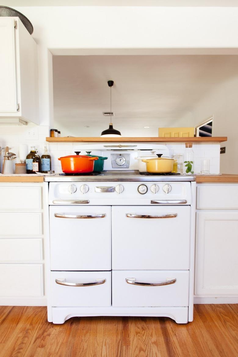 A white retro stove, with orange, yellow, and turquoise dutch ovens. White cabinets with wood counters.