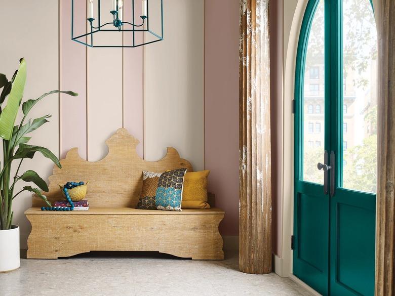 Wooden bench with multi-colored pillows in front of a baby pink wall next to a green-blue door