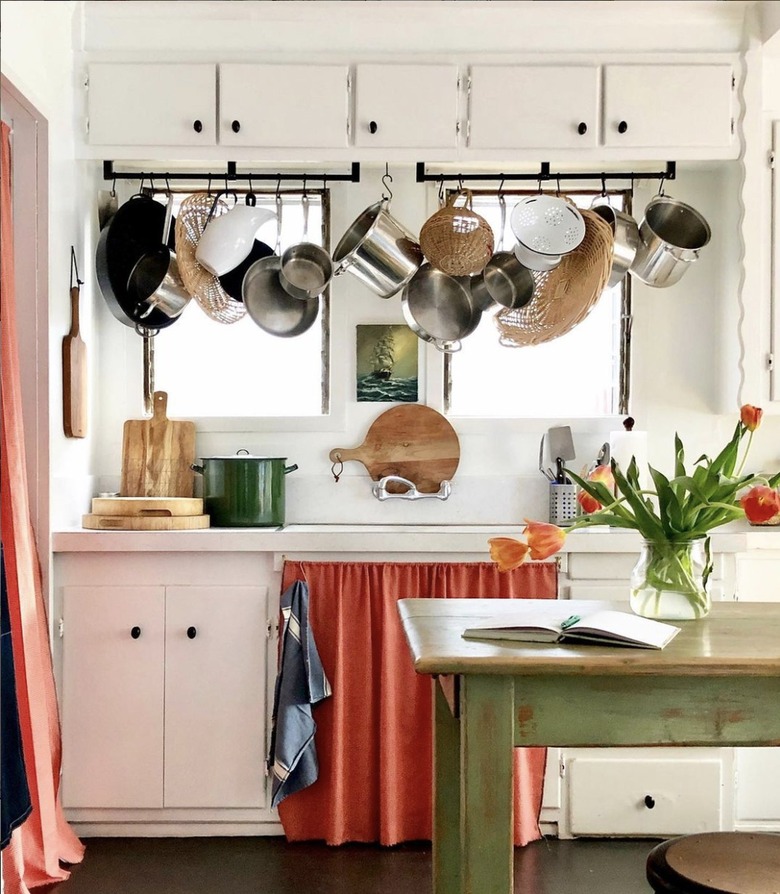 White kitchen with bright colored cabinet curtain and hanging pots.