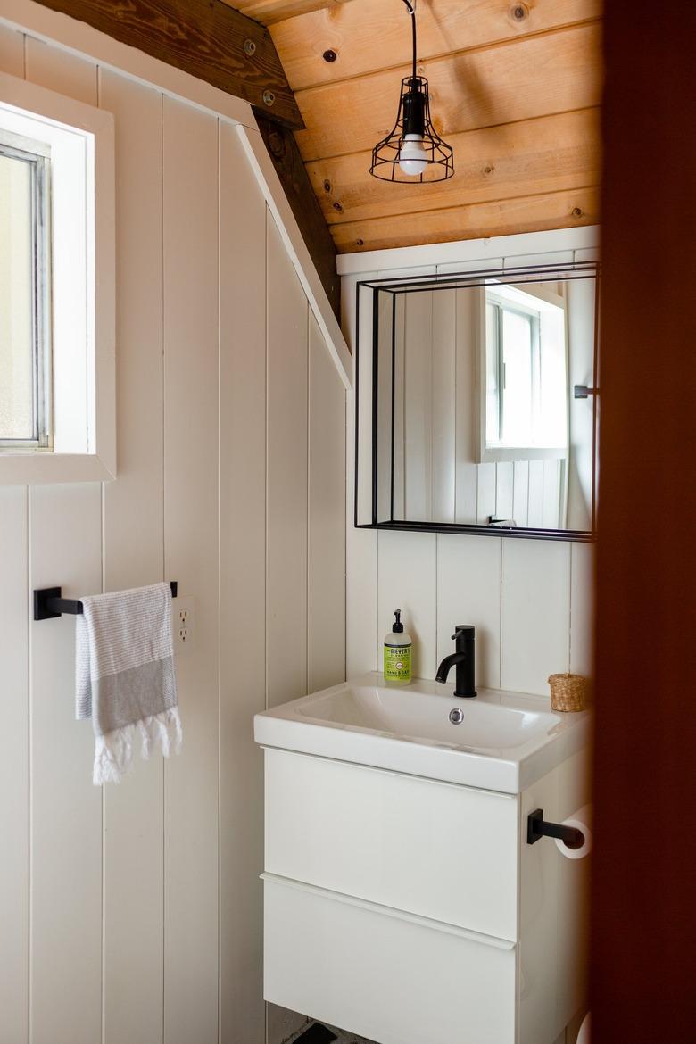 small modern bathroom with wood ceiling with mirror, small white sink, black faucet, and white hand towel