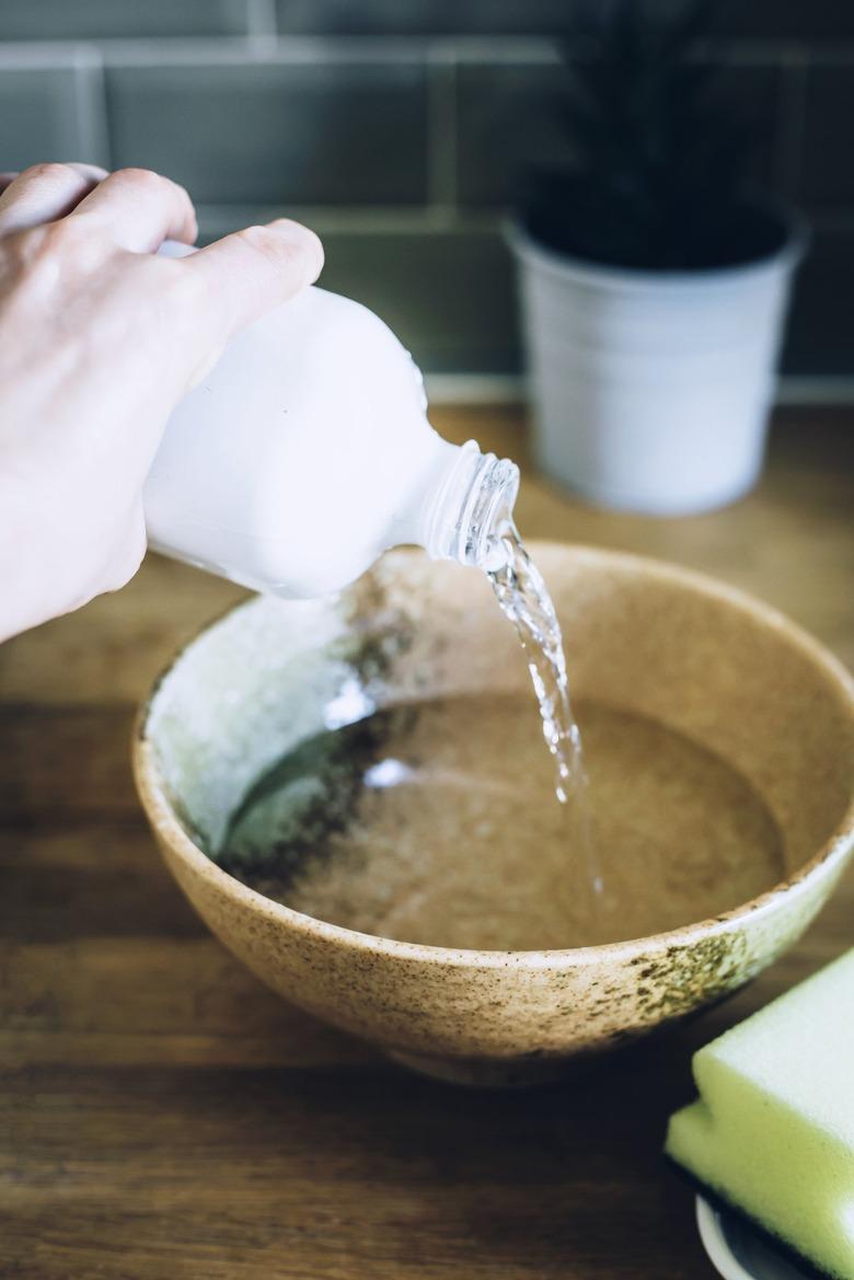 Hand pouring clear liquid into brown ceramic bowl on wood countertop against turquoise backsplash