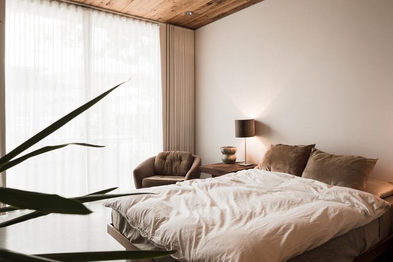 Minimalist bedroom with wood ceiling, beige curtains, brown accent chair, and neutral-beige accents.