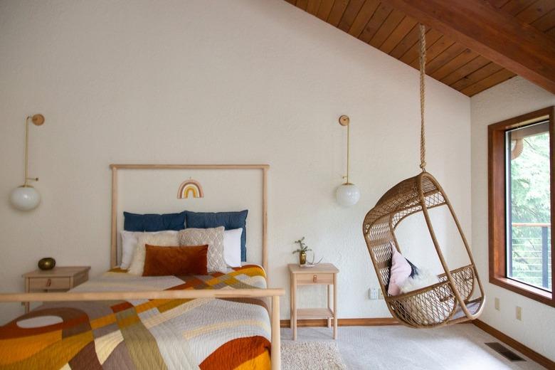 Bedroom with wood ceiling, casement windows. Neutral bedding, fiber rug, globe sconce lights, hanging boho chair.