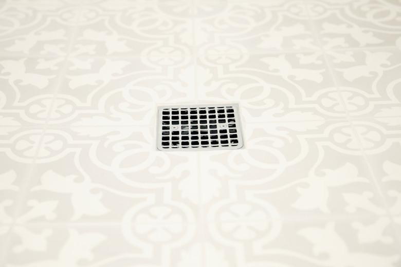 Closeup of white and grey patterned bathroom floor tiles with a chrome square grille shower drain in the middle.