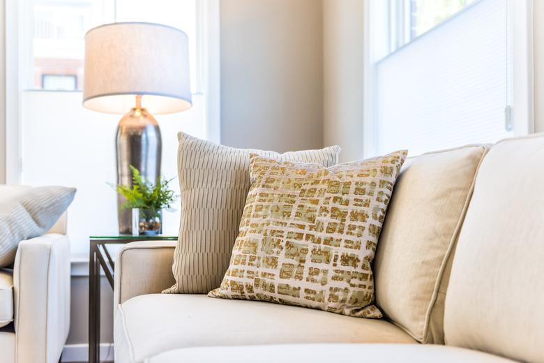 Closeup of two pillows on couch or sofa by bright window in modern apartment, house or home with staging of large beige, neutral white colors, lamp