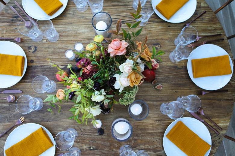 a rustic round wooden table with a large floral centerpiece and a gold napkin in the center of each plate