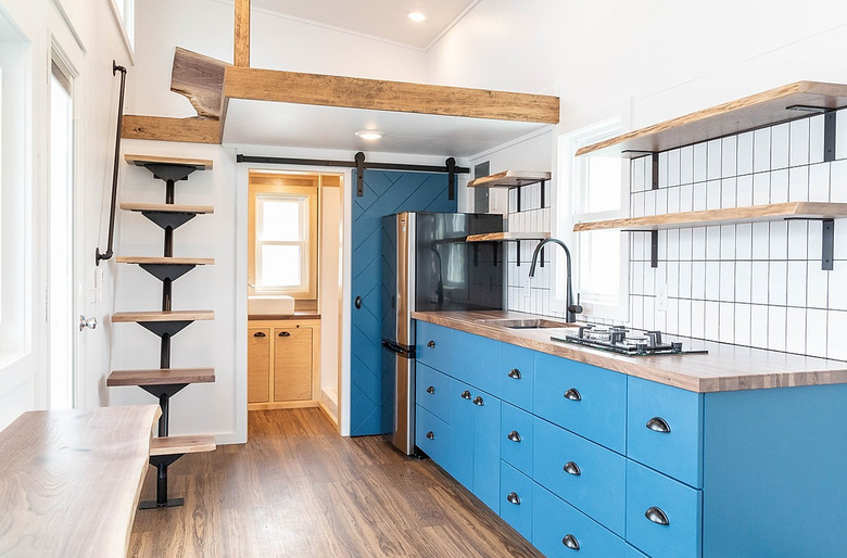 tiny house stairs in small home with blue cabinets, open wood shelves, black finish faucet and hardware, tile backsplash, hardwood floor, wood beams, wood and metal stairs.