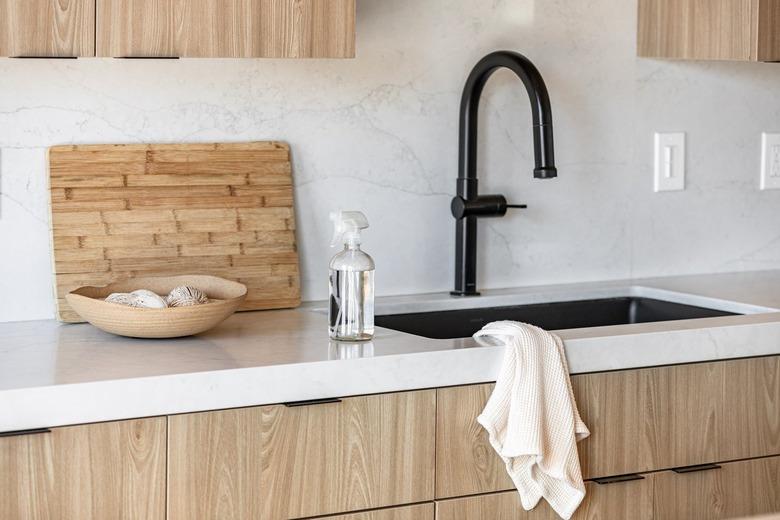 Spray bottle of cleaning liquid on kitchen counter with beige accent bowl and wood cutting board and black faucet sink