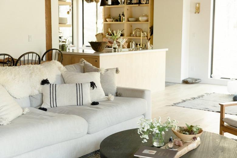 Living room with a white sofa with tassel accent pillows, round black coffee table. A kitchen with wood shelving and cabinets.