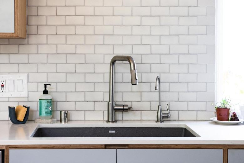 A stainless steel kitchen sink faucet in front of a white subway tile backsplash.