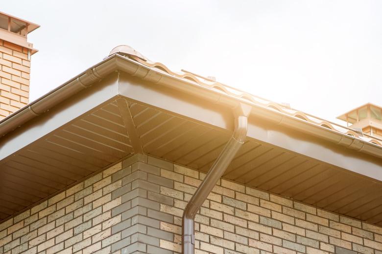 Corner of house with new brown metal tile roof and rain gutter. Metallic Guttering System, Guttering and Drainage Pipe Exterior