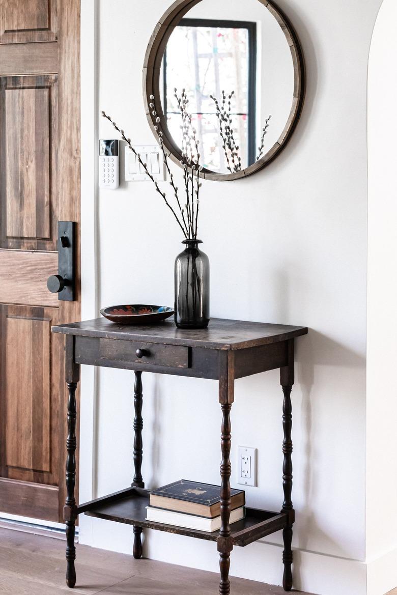 A side table with a black vase of willow cuttings and a round mirror and wood door