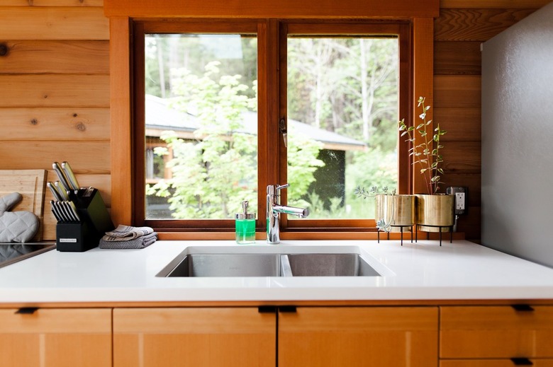 A white kitchen counter with a drop-in sink. Wood cabinets, walls, and window frame. Plants in gold planters.