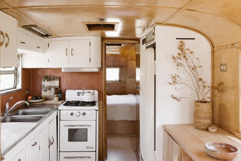 Interior of a camper with a white cabinet kitchen, Southwest rug, retro refrigerator, stools, wood table, basket vase with dried flowers.