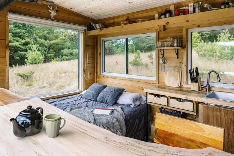 Interior of rustic wood-paneled room with bedding, kitchenette, and large windows against grassy field with trees