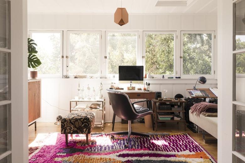 An sunny office with wood furniture, houseplants, colorful rug and geometric pendant light.