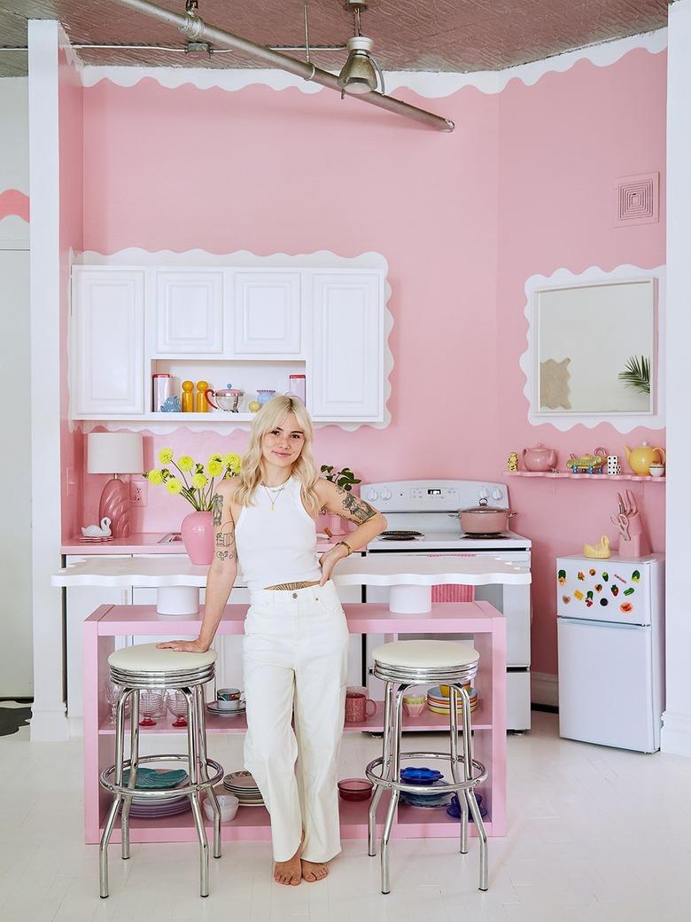 Furniture designer Sophie Collé standing in a predominately pink kitchen, wearing white jeans and a white tank top with shoulder-length blonde hair.