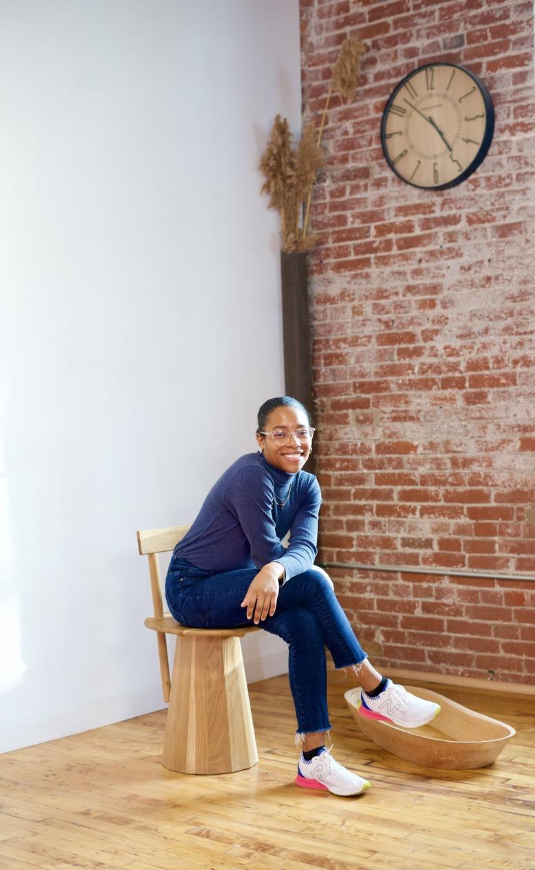 Furniture designer Tiarra Bell sitting on one of her wood chairs while wearing blue jeans, a long-sleeve blue top, glasses, and white sneakers.