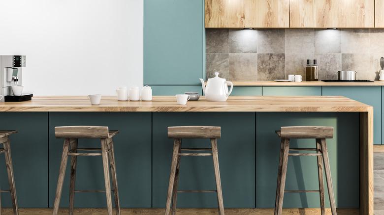 modern kitchen with dark blue-green lower and full-height cabinets, light wood countertops, light wood upper cabinets, natural wood bar stools, and a oversized square tile stone backsplash