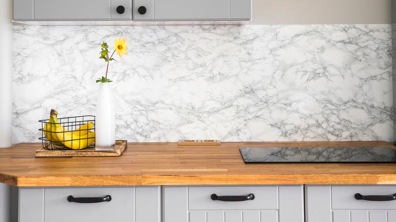 kitchen with gray cabinets, marble backplash, wood countertops, and black hardware with a basket of bananas and a vase containing a yellow flower on the counter