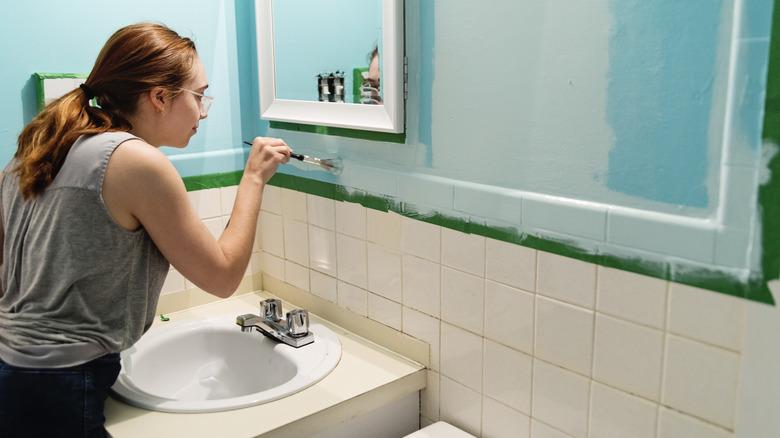 a woman is repainting a bathroom and has painters' tape up over white tiles.