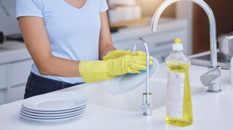 A woman is shown from the shoulders-down wearing yellow cleaning gloves and washing dishes at the kitchen sink.