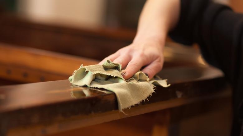 A hand wiping down a piece of wooden furniture with a green cloth.