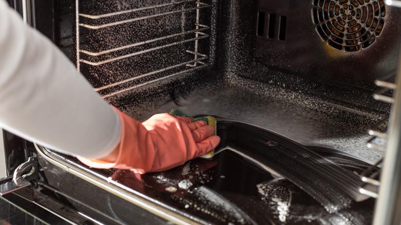 A gloved hand cleaning the inside of an oven with a sponge.