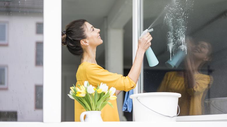 A woman in a yellow top is spraying cleaning solution onto a large glass window.