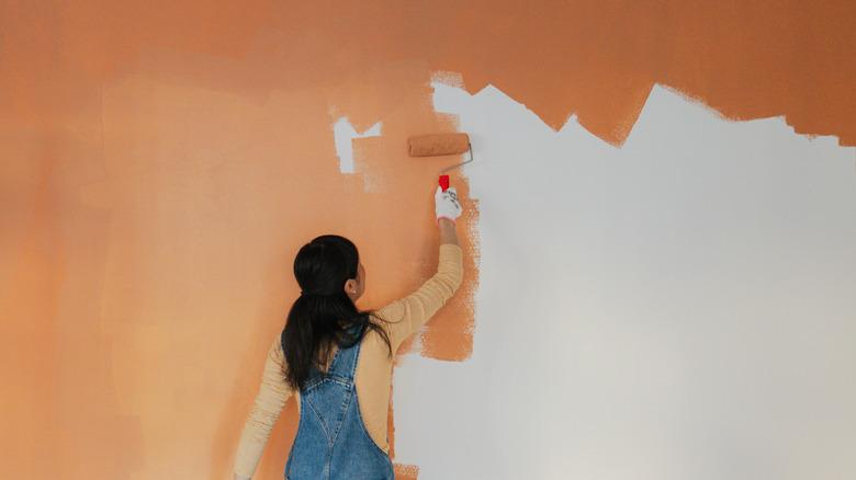 woman painting wall with orange paint wearing orange shirt and blue jean overalls