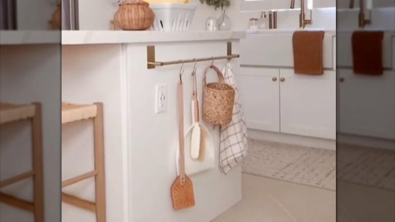 brass towel rod installed as a kitchen rail storage system on the side of the island with S hooks holding a towel, basket, dustpan, and leather fly swatter