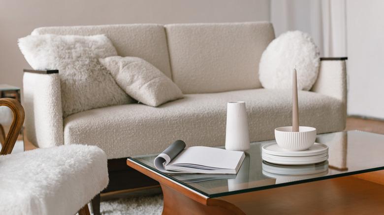 A neutral living room with a white boucle sofa, white faux-fur chair, white pillows, and white decor on a wood and glass coffee table