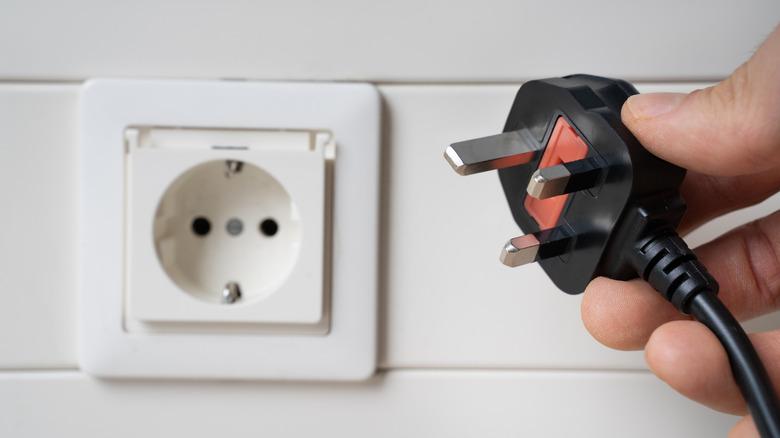 A hand holds an electrical plug in the foreground, with a different type of outlet in the background.