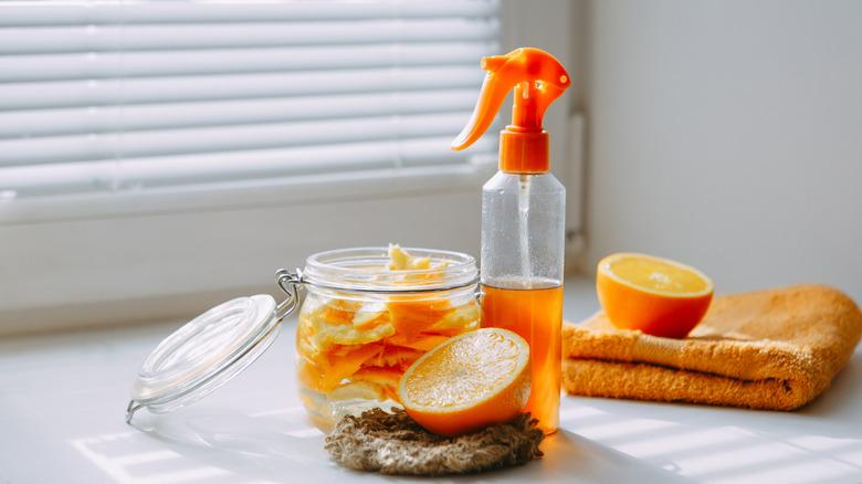 citrus peels in a jar next to a spray bottle of orange liquid and half an orange on a sponge with the other half of the orange on an orange towel, everything sitting together on a white table in front of white blinds