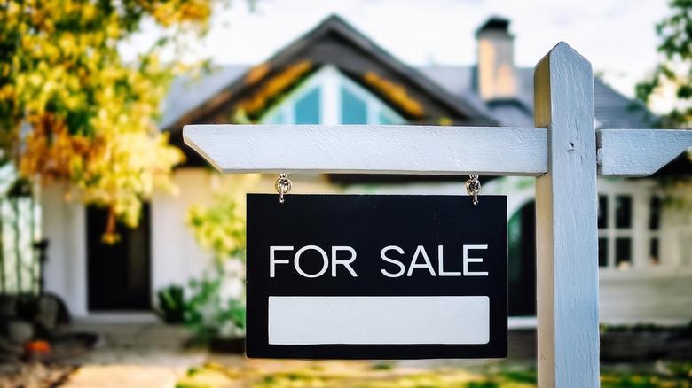 The image is focused on a black and white For Sale sign, with a blurred home in the background.